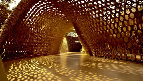 A low-angle view inside a large arched tunnel with a honeycomb lattice wall structure casting strong dappled shadows on the floor.