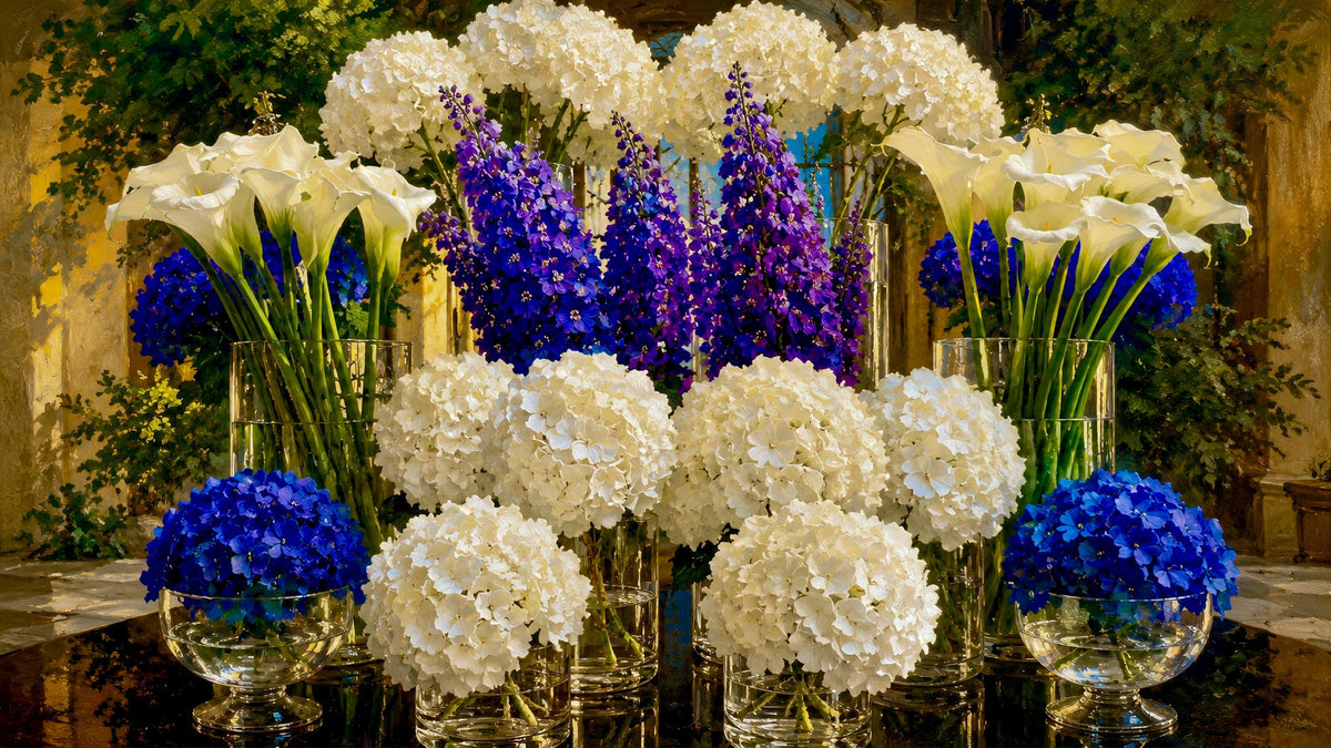 A floral arrangement of white calla lilies, white hydrangeas, and purple delphiniums in clear glass vases and bowls, set against a warm, softly blurred background.